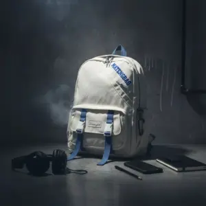 Cream white canvas backpack with blue webbing straps, front buckle pockets, and a 'MATENGBAG' logo, styled in a moody setting with headphones and a notebook.
