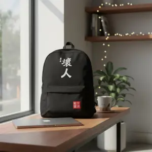 Black canvas backpack with bold white Japanese or Chinese graphic text and a small red tag, sitting on a wooden desk next to a silver laptop and coffee mug in a sunlit room.