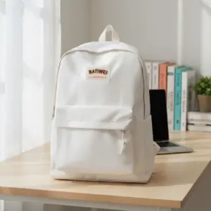 BATIWEI White Minimalist Backpack with front pocket and orange logo patch, sitting on a wooden desk next to a laptop and books in a brightly lit room.