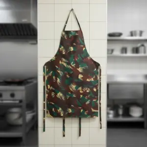 Camouflage print kitchen apron in military green, brown, and tan hanging on a white tiled wall in a professional kitchen setting.