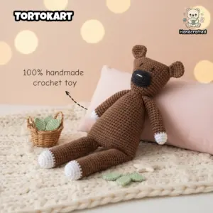 100% handmade brown crochet teddy bear inspired by Mr. Bean’s Teddy, featuring white paws and a black muzzle, resting against a pink pillow on a cream chunky knit blanket.