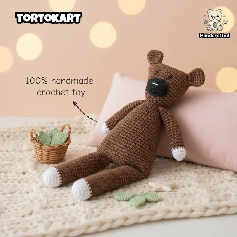 100% handmade brown crochet teddy bear inspired by Mr. Bean’s Teddy, featuring white paws and a black muzzle, resting against a pink pillow on a cream chunky knit blanket.