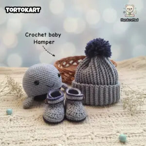 Handmade crochet baby gift hamper featuring a grey amigurumi whale plushie, a ribbed baby beanie with a navy pom-pom, and matching crochet booties on a textured cream blanket.