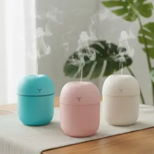 Three small, rounded mini humidifiers in teal, pink, and white colors emitting a steady stream of cool mist on a wooden table with a leafy green plant in the background.