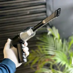 A hand in a white glove holding a black 3-in-1 portable wireless vacuum cleaner and blower with a long brush attachment, shown against a dark textured background with a green plant.