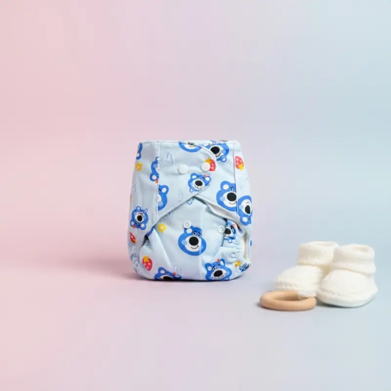 Light blue reusable cloth diaper with a cartoon bear print, featuring adjustable white snap buttons, shown next to white knitted baby booties and a wooden teething ring.