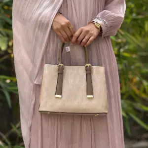 A high-quality women's luxury handbag in a metallic mauve-bronze with a vertical pleated texture and gold hardware, held by a woman wearing a matching pleated mauve dress and gold jewelry.