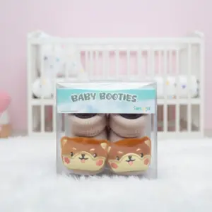 Tan knit baby booties with cute embroidered Shiba Inu dog faces on the toes, displayed in clear Semega brand packaging.