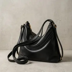 The file 1 (20).webp displays a large, slouchy black leather ladies sling bag resting on a textured light grey surface against a matching concrete wall background, highlighting its gold-toned zippers, hardware accents, and elegant layered pleat detailing across the front curve.