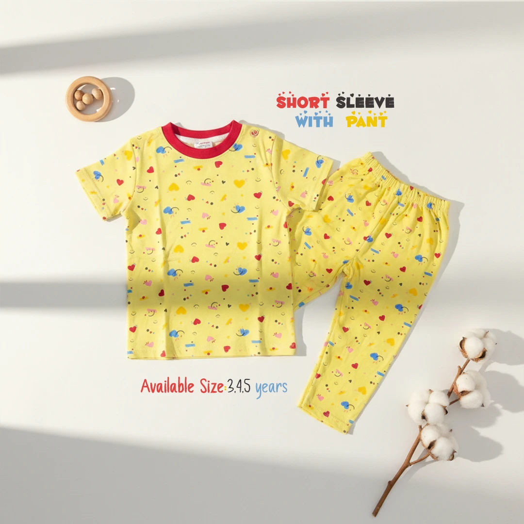 The image displays a bright yellow toddler lounge set patterned with colorful hearts, stars, and smiley faces, including a short sleeve round neck t-shirt with a red collar trim and matching long pants, laid flat on a white surface with shadows next to a wooden rattle and a cotton branch.
