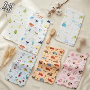 The photos displays six rectangular muslin baby burp cloths arranged neatly on a light beige fabric backdrop. The collection includes a white cloth with an avocado and banana print, a white cloth with a transportation and car print, a white windowpane check cloth with whale graphics, a light blue cloth with space rockets and flying saucers, an orange cloth patterned with racing cars, and a pink cloth featuring cartoon characters and flowers. A wooden baby brush, a crochet rattle, and cotton stems decorate the flat lay layout.