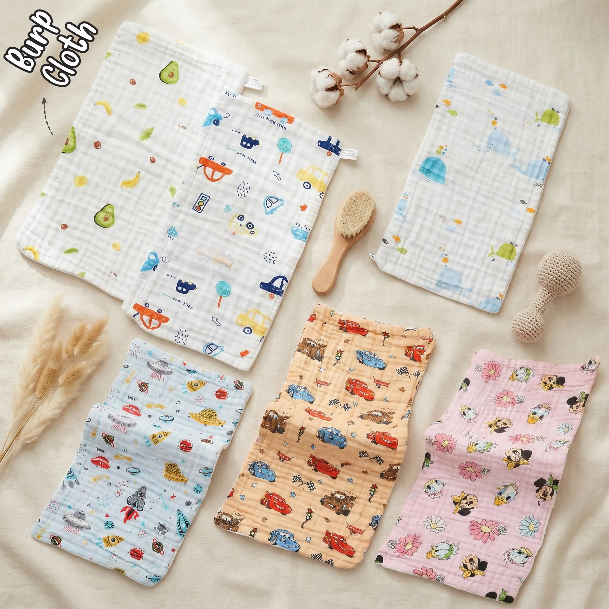 The photos displays six rectangular muslin baby burp cloths arranged neatly on a light beige fabric backdrop. The collection includes a white cloth with an avocado and banana print, a white cloth with a transportation and car print, a white windowpane check cloth with whale graphics, a light blue cloth with space rockets and flying saucers, an orange cloth patterned with racing cars, and a pink cloth featuring cartoon characters and flowers. A wooden baby brush, a crochet rattle, and cotton stems decorate the flat lay layout.