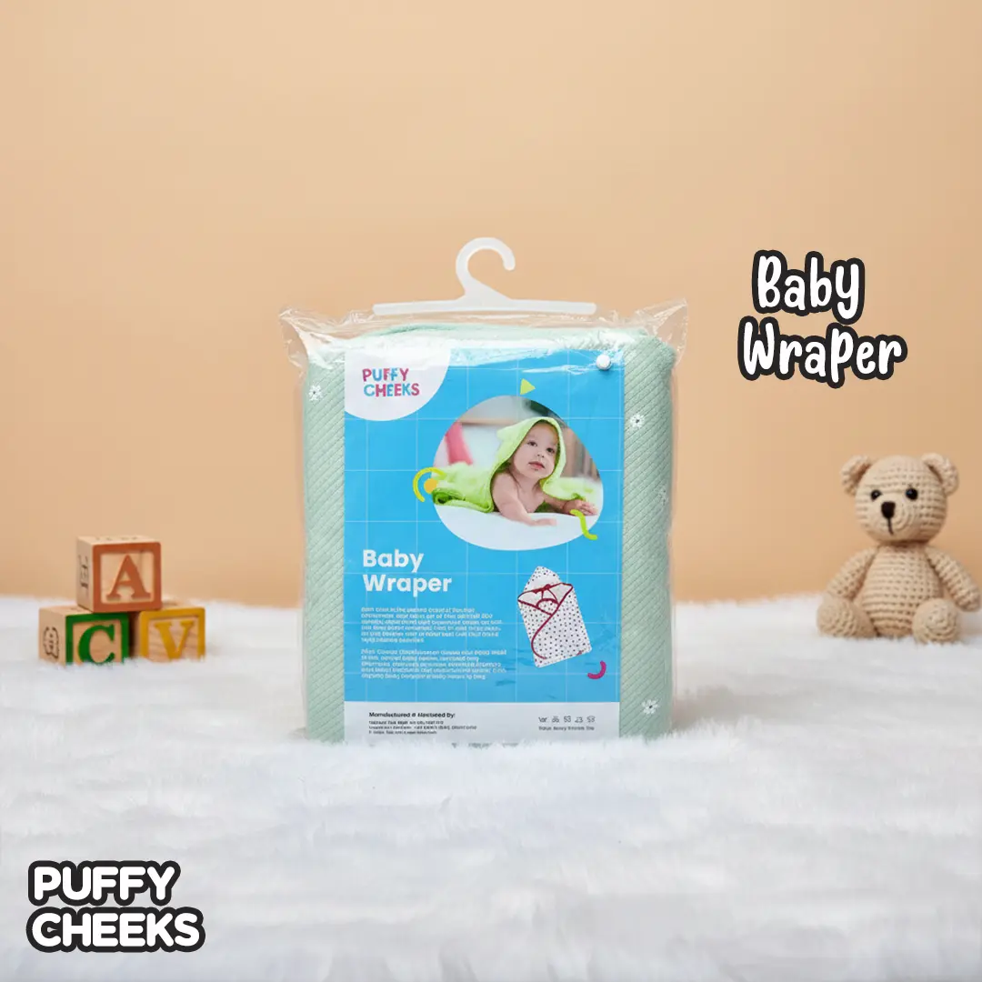 The image displays a retail-packaged Puffy Cheeks Baby Wrapper in mint green with white floral accents, enclosed in a transparent plastic pouch with a white hanger. The blue product label features a circular insert of a baby wearing a hooded blanket, positioned on a white furry surface next to wooden alphabet blocks and a crochet teddy bear.