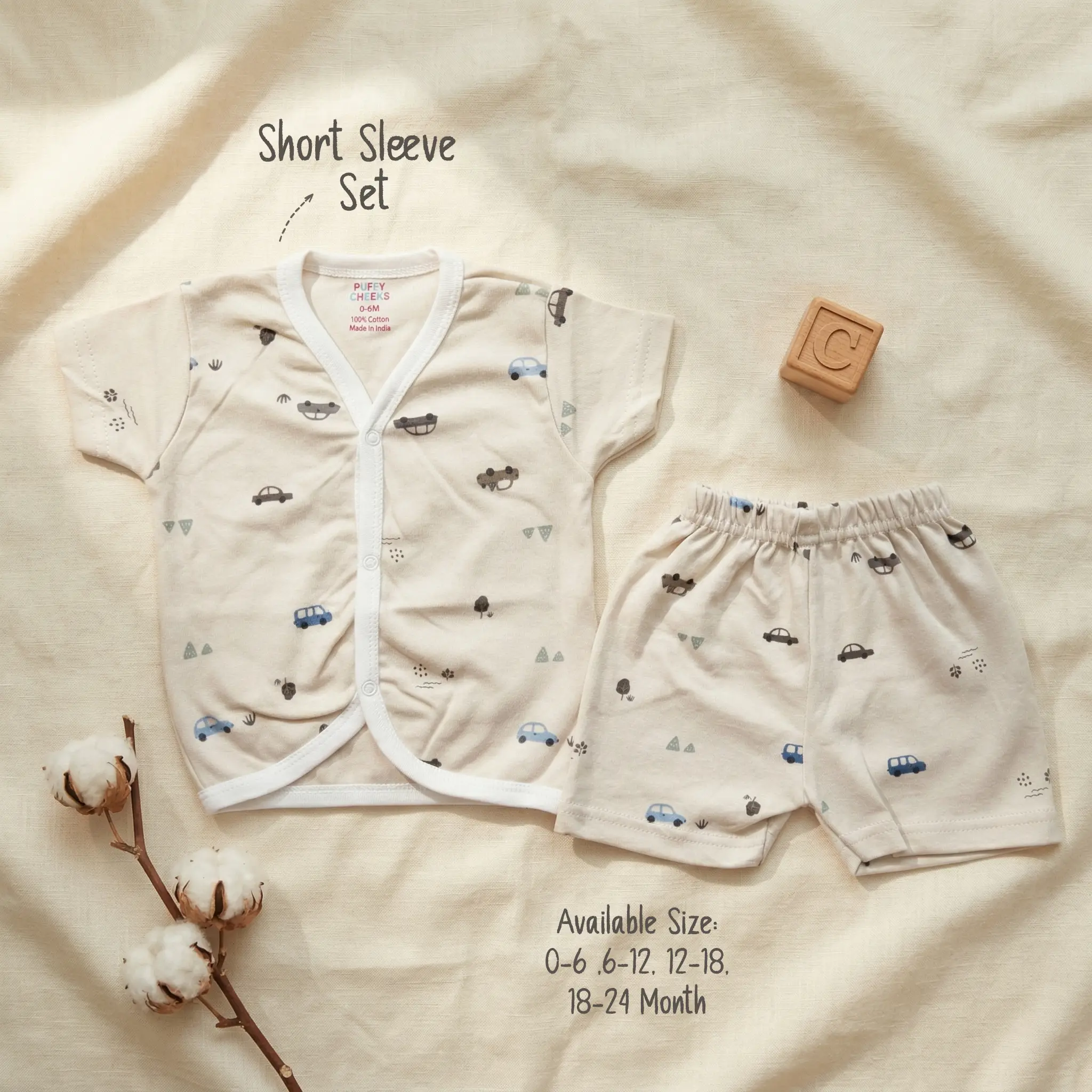 Infant short sleeve front-open buttoned shirt and matching shorts set by Puffy Cheeks flat-laid on a cream textured fabric, featuring a minimalist repeating print of small blue, grey, and brown cars interspersed with tiny trees and dots on beige cotton fabric, alongside a wooden alphabet block with the letter C and a natural raw cotton boll branch.