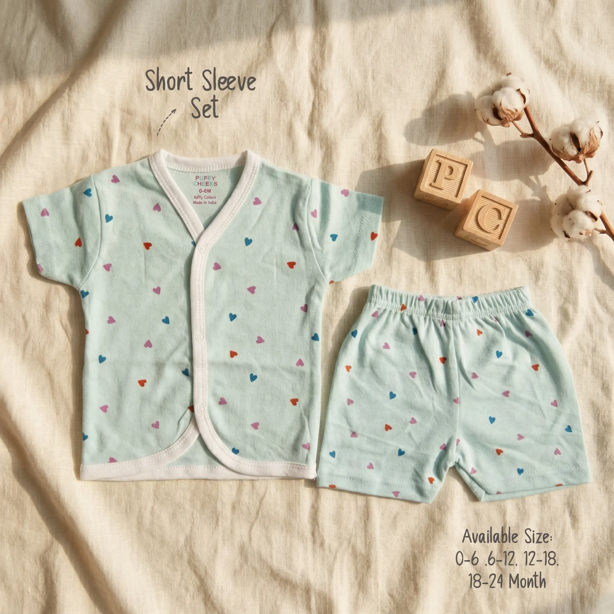 Infant short sleeve front-open buttoned shirt and matching shorts set by Puffy Cheeks flat-laid on a cream textured fabric, featuring a minimalist repeating tiny heart print in blue, purple, and red scattered over a light mint green cotton fabric, finished with white bound trims, positioned beside wooden alphabet blocks showing the letters P and C and a branch of raw natural cotton bolls.