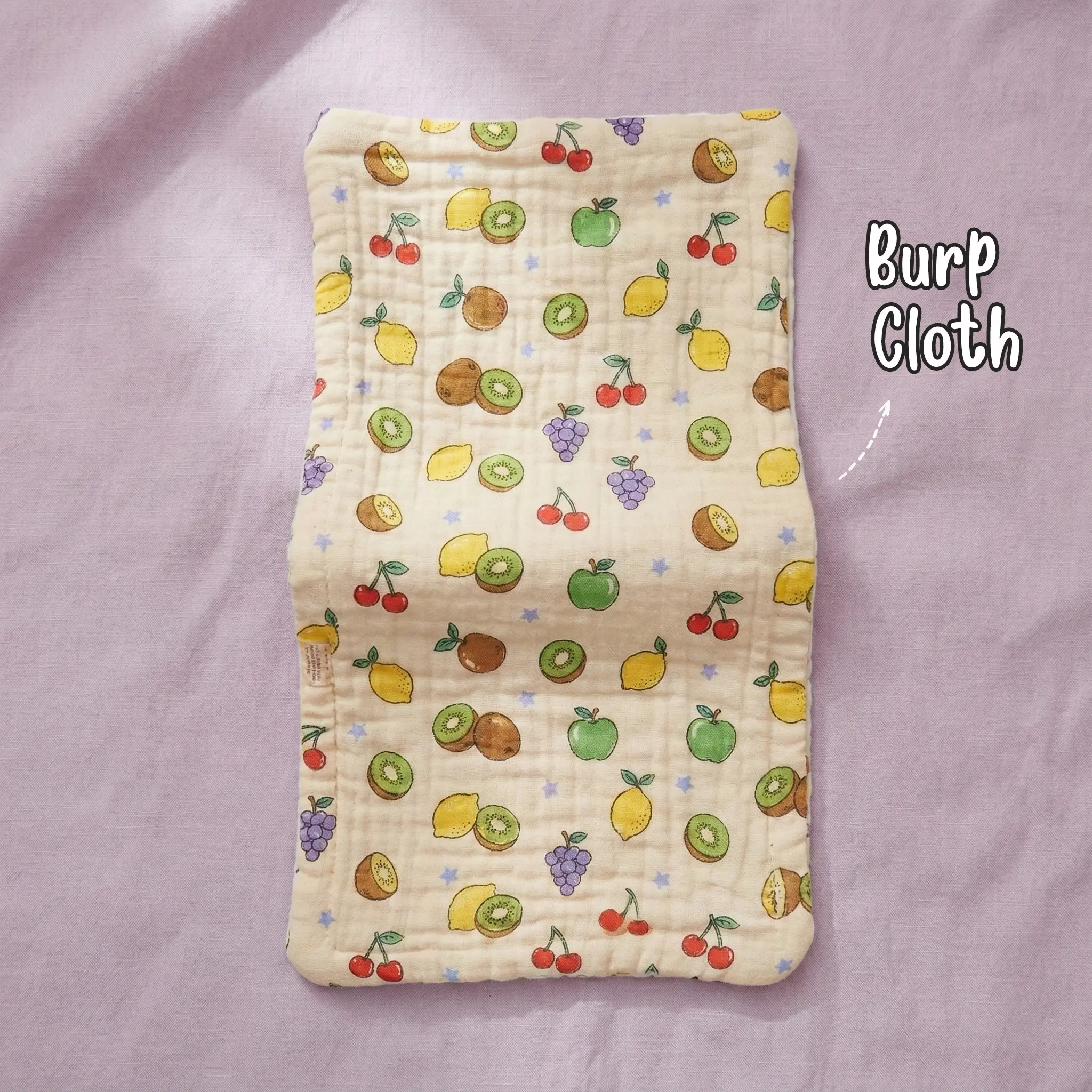 The picture shows a single rectangular light orange or peach-colored muslin baby burp cloth by Puffy Cheeks with a crinkled texture, patterned with an all-over racing car print featuring red, blue, and brown cartoon cars, checkered racing flags, and tiny traffic lights, laid flat on a soft purple fabric surface.