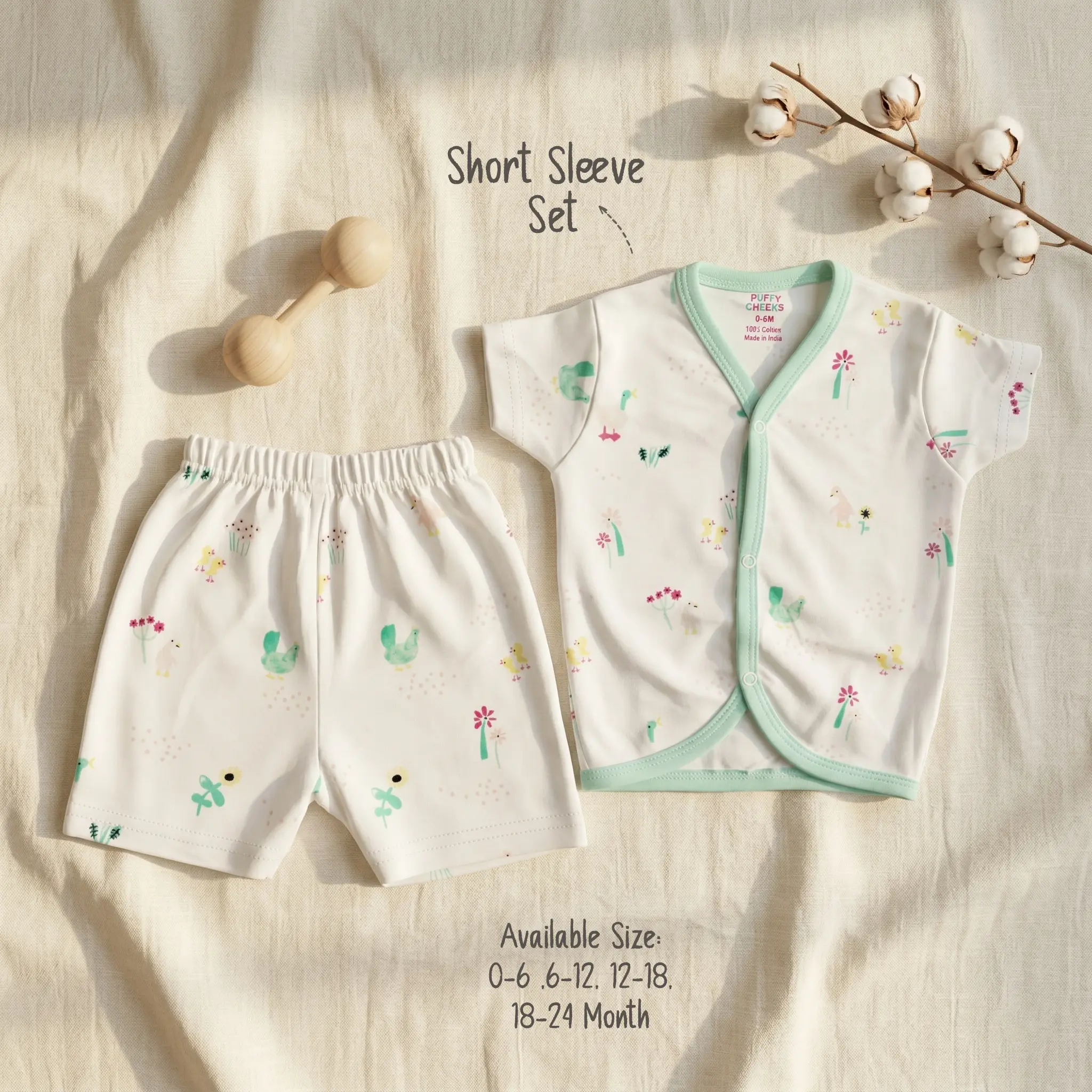 Infant short sleeve front-open buttoned shirt and matching shorts set by Puffy Cheeks flat-laid on a cream textured fabric, featuring an all-over garden pond illustration print with green ducks, yellow chicks, pink flowers, and reeds on white cotton fabric, trimmed with a mint green bound V-neckline border, alongside a wooden dumbbell rattle and a natural raw cotton boll branch.