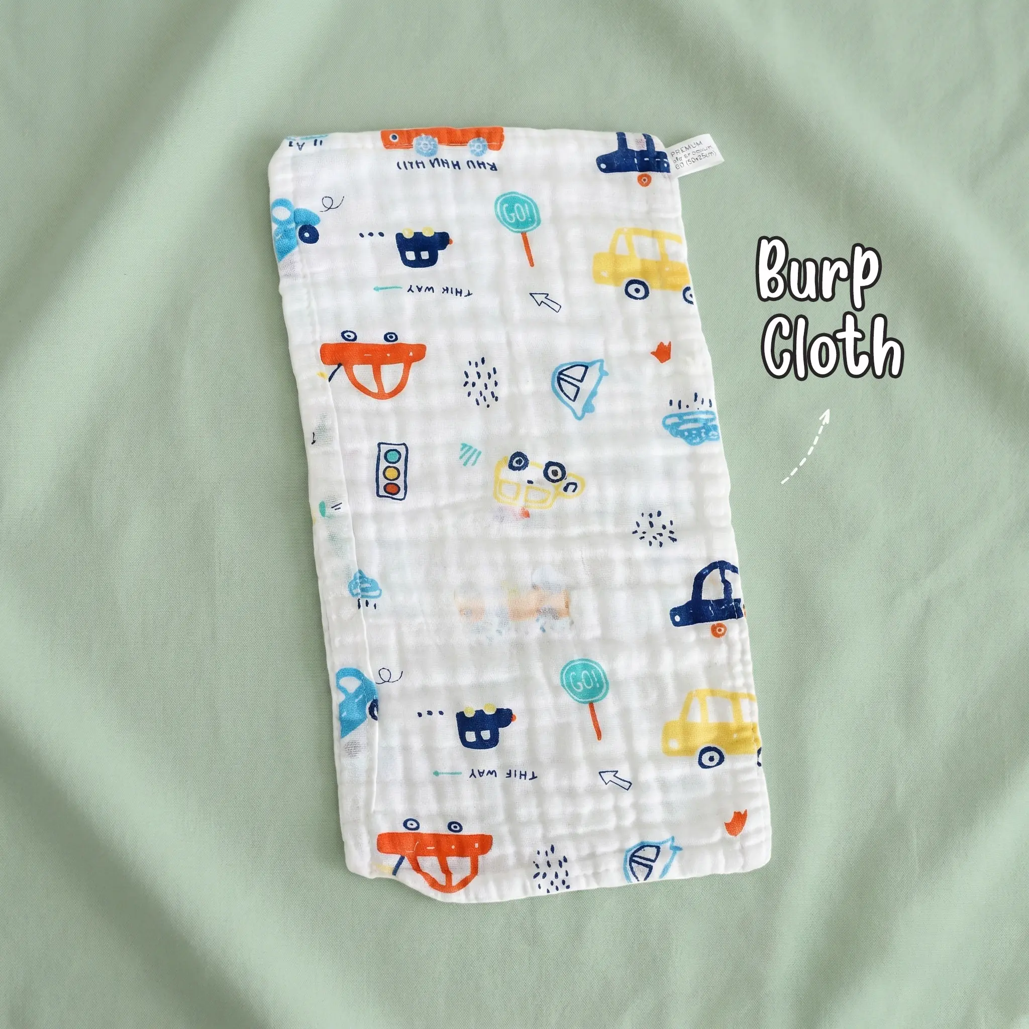 The photo shows a single rectangular white muslin baby burp cloth by Puffy Cheeks with a crinkled texture, decorated with a transportation print of colorful cars, buses, traffic lights, and "GO!" signs, laid flat on a smooth mint green fabric surface.