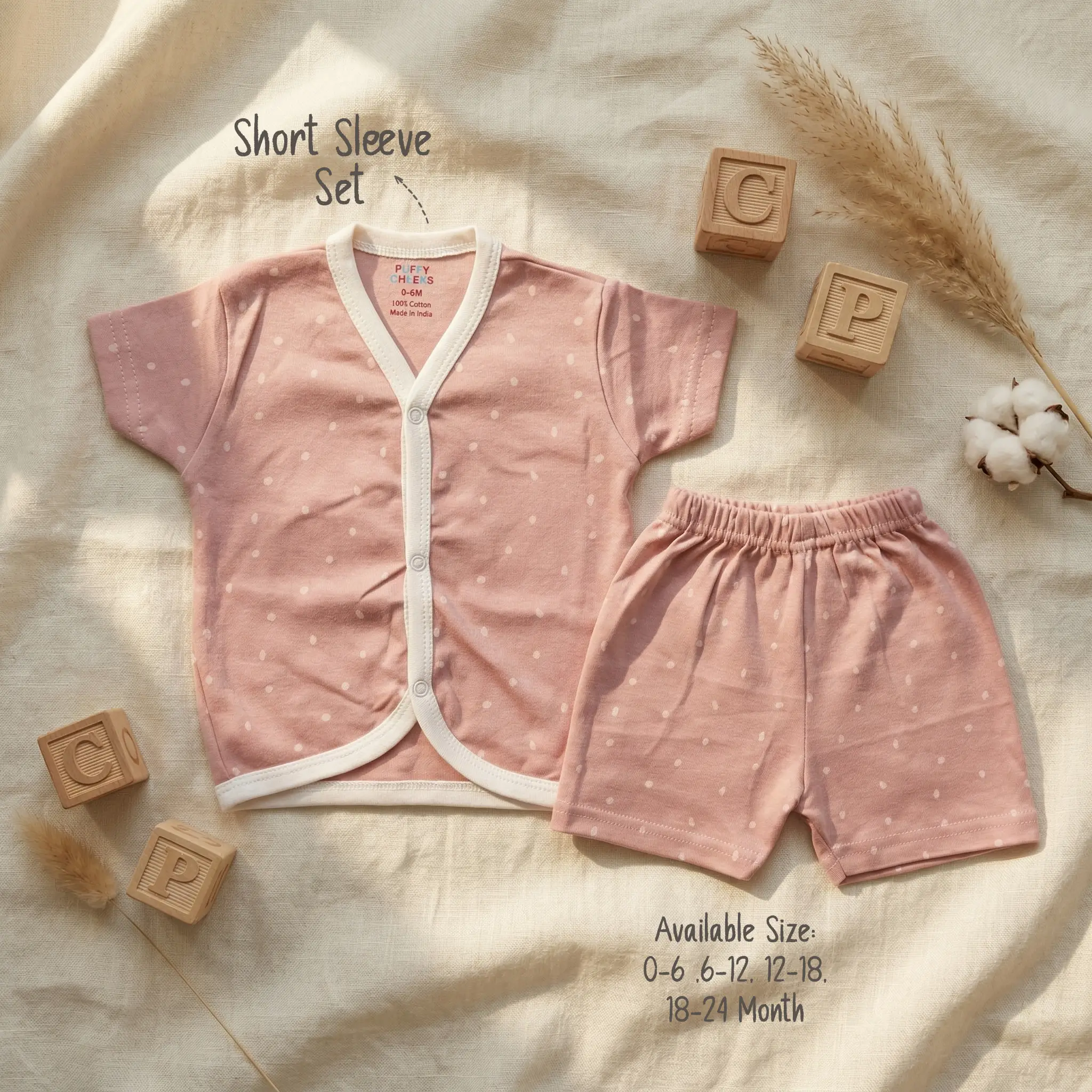 Infant short sleeve front-open buttoned shirt and matching shorts set by Puffy Cheeks flat-laid on a cream textured fabric, featuring a subtle all-over pattern of small white polka dots on dusty rose pink cotton fabric, surrounded by wooden alphabet blocks with letters C and P, a single dried grass stalk, and a natural raw cotton boll branch.