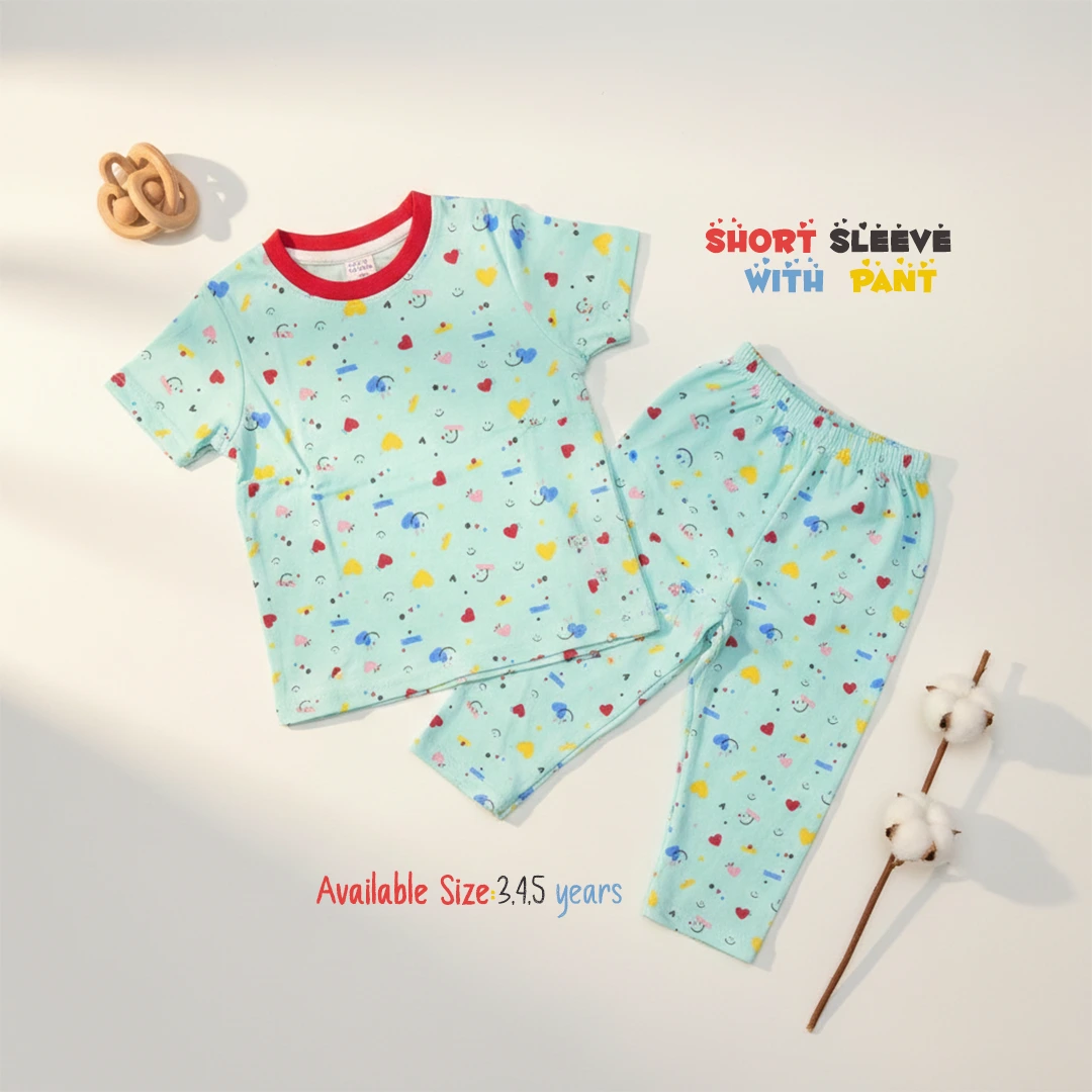 The image displays a mint green toddler lounge set patterned with colorful hearts, stars, and smiley faces, including a short sleeve round neck t-shirt with a red collar trim and matching long pants, laid flat on a white surface next to a wooden rattle and a cotton branch.