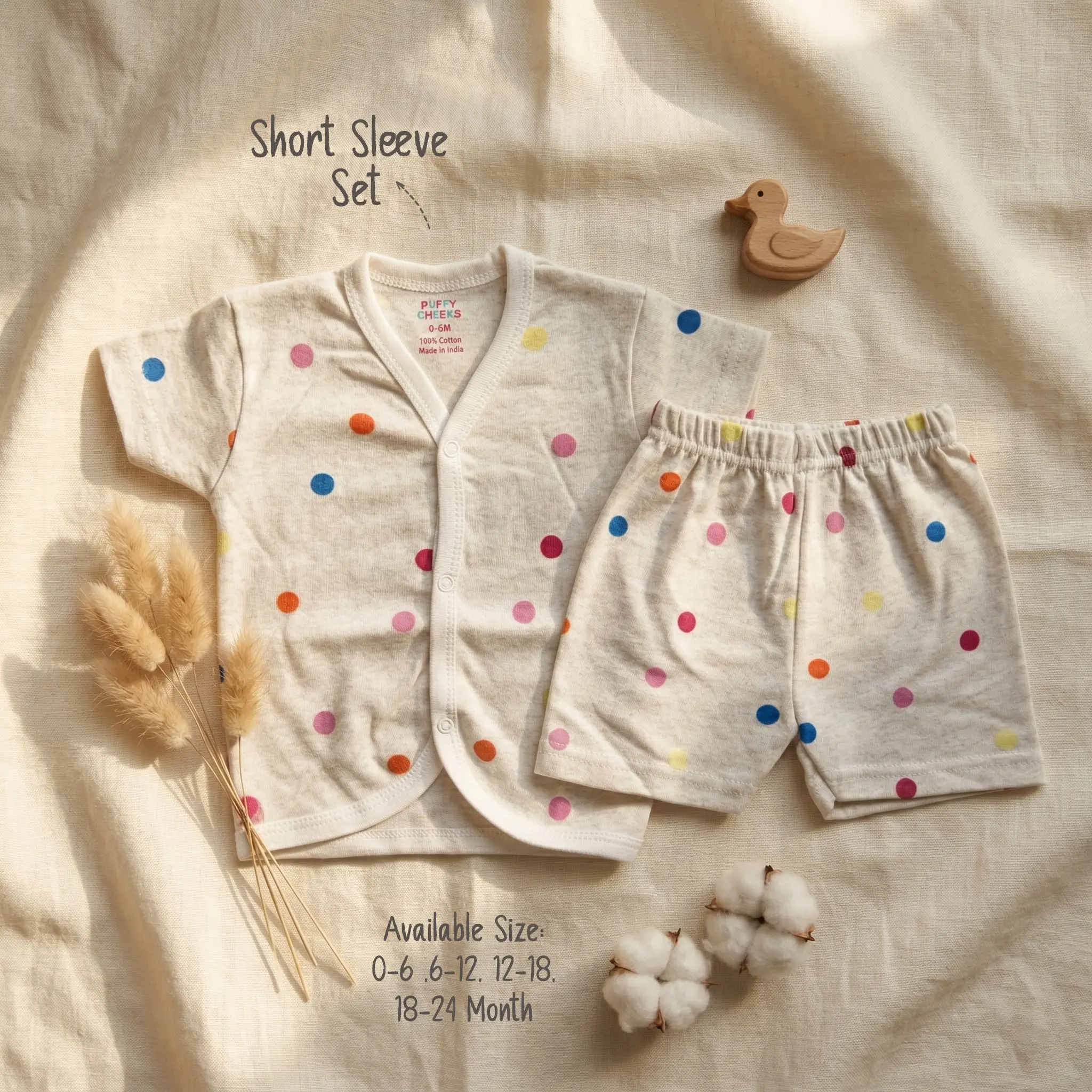 Infant short sleeve front-open buttoned shirt and matching shorts set by Puffy Cheeks flat-laid on a cream textured fabric, featuring a playful scattered multi-colored polka dot print in blue, orange, pink, yellow, and red on a cream-heather cotton fabric, styled next to a small wooden duck figurine, dried grass stalks, and two natural raw cotton bolls.
