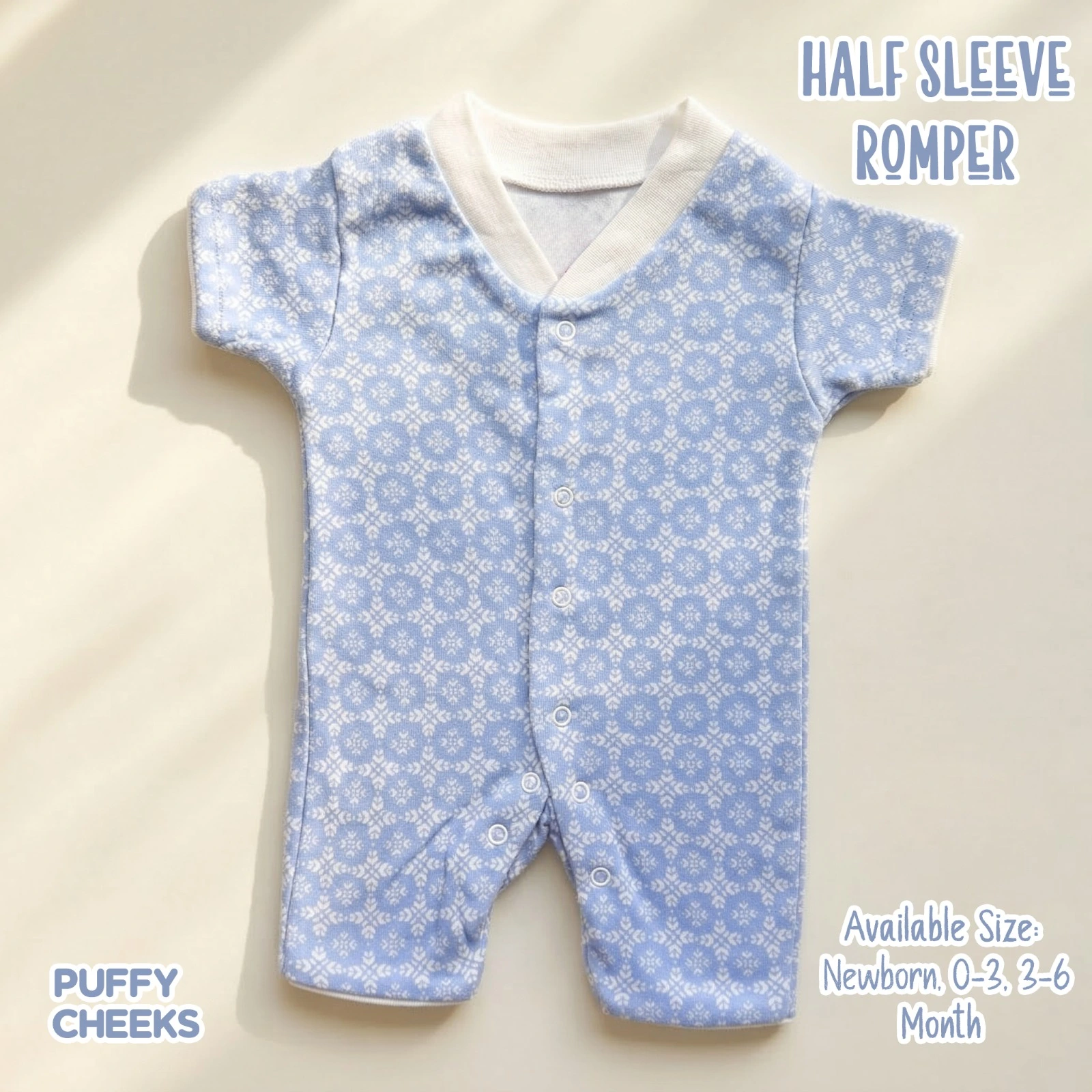 THE IMAGE shows a light blue, half-sleeve baby romper by Puffy Cheeks featuring an all-over white geometric snowflake pattern. The garment is designed with a soft white v-neck style ribbed collar, short sleeves, a full snap-front button closure running down the center, and snap buttons along the crotch inseam, flat-laid on a clean cream surface under soft lighting.