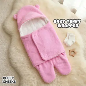 The image shows a plush pink baby terry wrapper by Puffy Cheeks with a hooded top featuring small bear ears and a split-leg or footed bottom design. The interior is lined with a soft, fluffy white fleece material, and the front has a wrap-around swaddle flap closure. It is flat-laid on a white faux fur rug next to a pair of white infant socks and a small star-shaped wooden teething toy.
