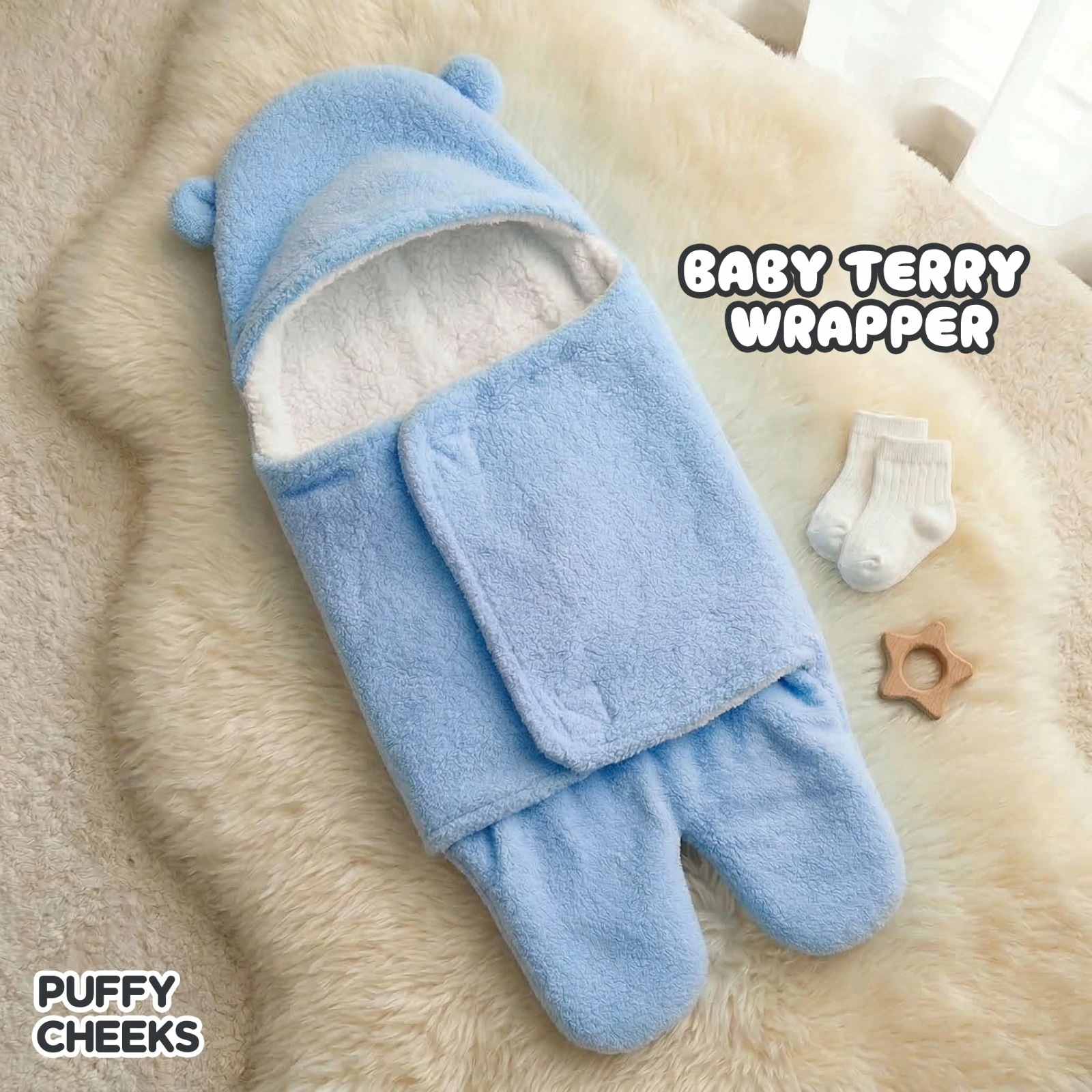 The image shows a plush light blue baby terry wrapper by Puffy Cheeks with a hooded top featuring small bear ears and a split-leg or footed bottom design. The interior is lined with a soft, fluffy white fleece material, and the front has a wrap-around swaddle flap closure. It is flat-laid on a white faux fur rug next to a pair of white infant socks and a small star-shaped wooden teething toy.