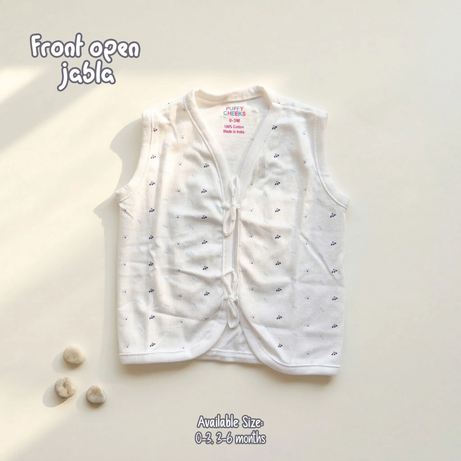 The image named WhatsApp Image 2026-06-17 at 12.51.45 PM (2).webp shows a sleeveless, front-open baby jabla by Puffy Cheeks featuring an all-over tiny, scattered dark geometric or floral print on a white base. The garment is designed with a v-neckline, soft white fabric trim along all borders, and two fabric tie-front closures at the center, flat-laid on a clean cream surface next to three small decorative stones.