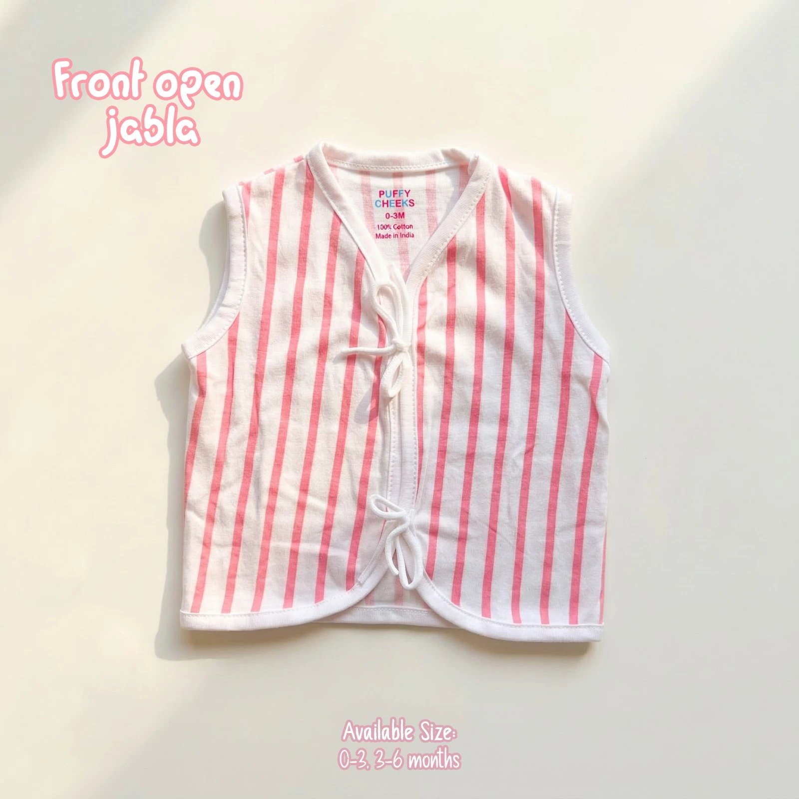 The image shows a white and pink vertically striped sleeveless front-open baby jabla by Puffy Cheeks. The garment features a v-neckline, soft white fabric trim lining all borders, and two fabric tie-front closures at the center, flat-laid on a smooth cream surface.