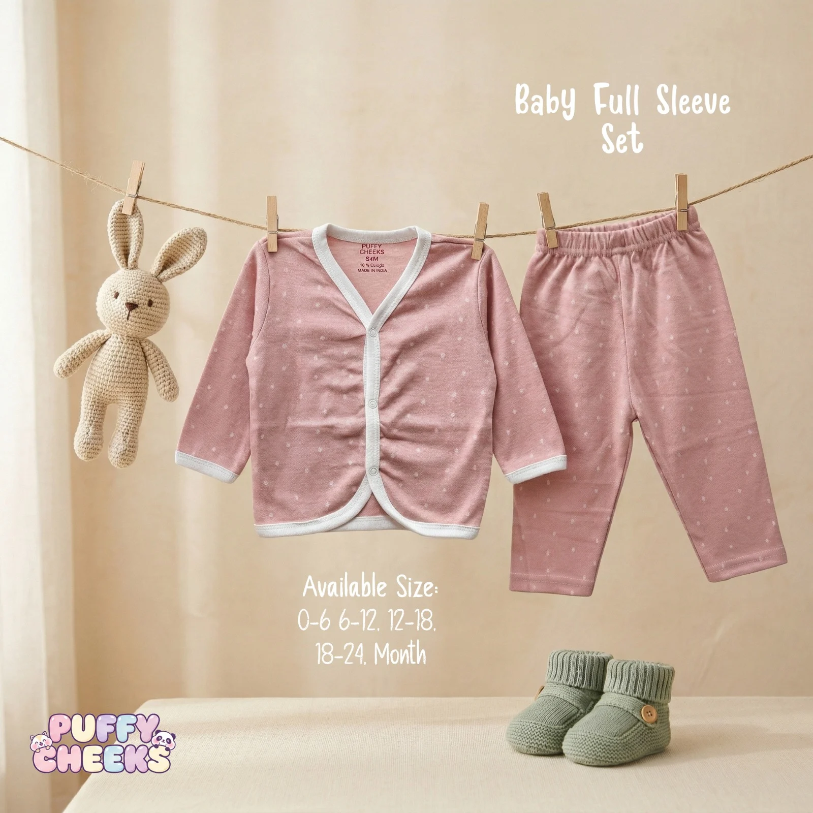 Baby full sleeve front-open top and matching pants set by Puffy Cheeks featuring a faint all-over white dot print on a dusty pink cotton fabric, with the long-sleeve cardigan top showing a white ribbed v-neck trim and snap-button closure hanging from a clothesline with wooden pegs next to matching elastic-waist pants and a small knit bunny toy, displayed above a pair of sage green knit infant booties on a soft surface.