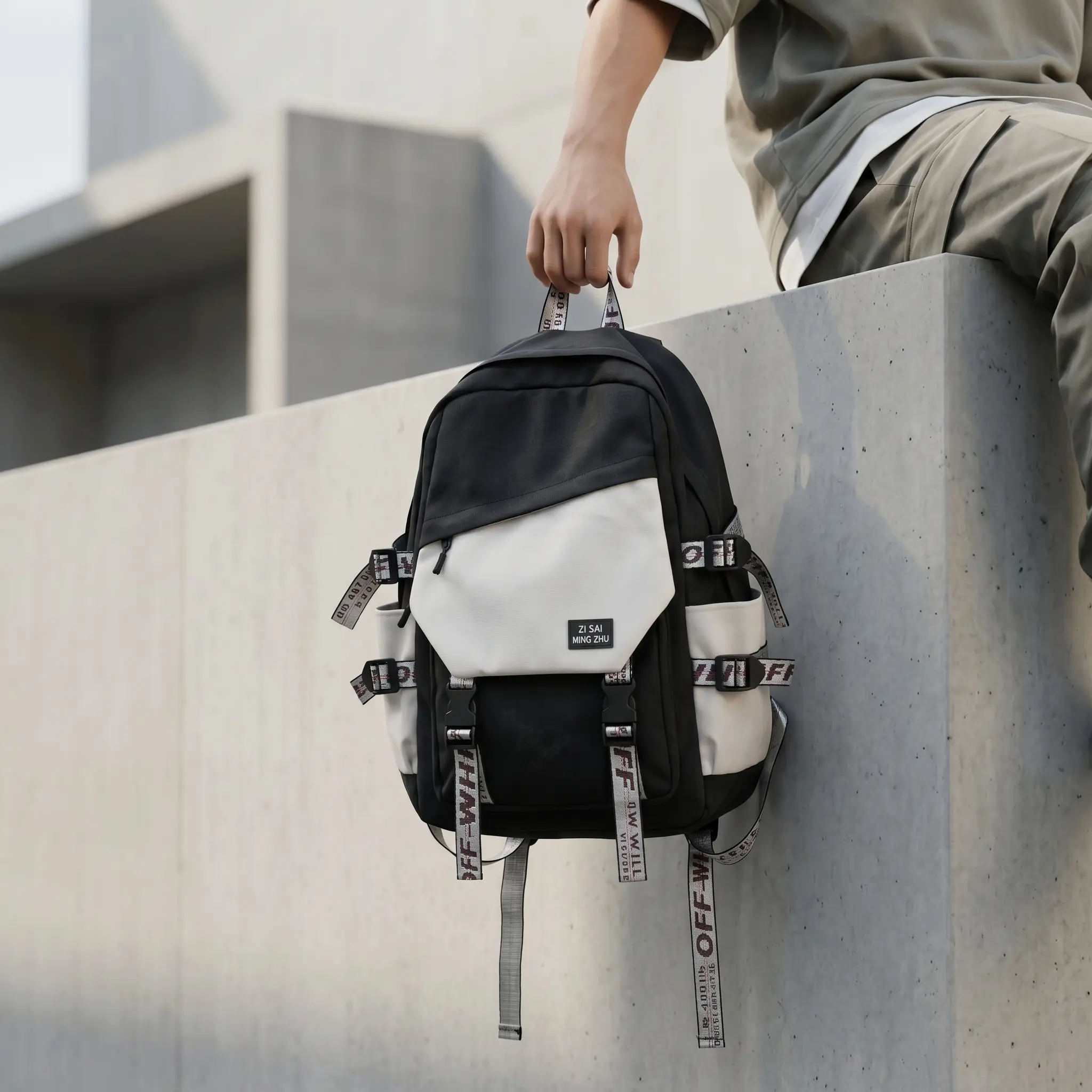 A person wearing an olive green jacket sitting on a modern concrete ledge, holding a black and off-white Tortokart urban utility backpack by its top webbed handle. The backpack features an asymmetrical white front zip pocket with a "ZI S.M MING ZHU" patch, industrial buckle straps with typography webbing, dual front clip straps, and cream side bottle pockets in an urban architecture setting.