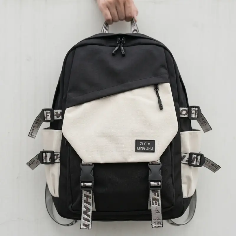 A person holding a durable black and beige contrast urban streetwear backpack by the top handle against a minimalist grey wall. The backpack features an asymmetric beige front zip pocket with a "ZI S.M MING ZHU" woven patch, side compression buckle straps with patterned industrial webbing, dual bottom clip straps, and cream side water bottle pockets.