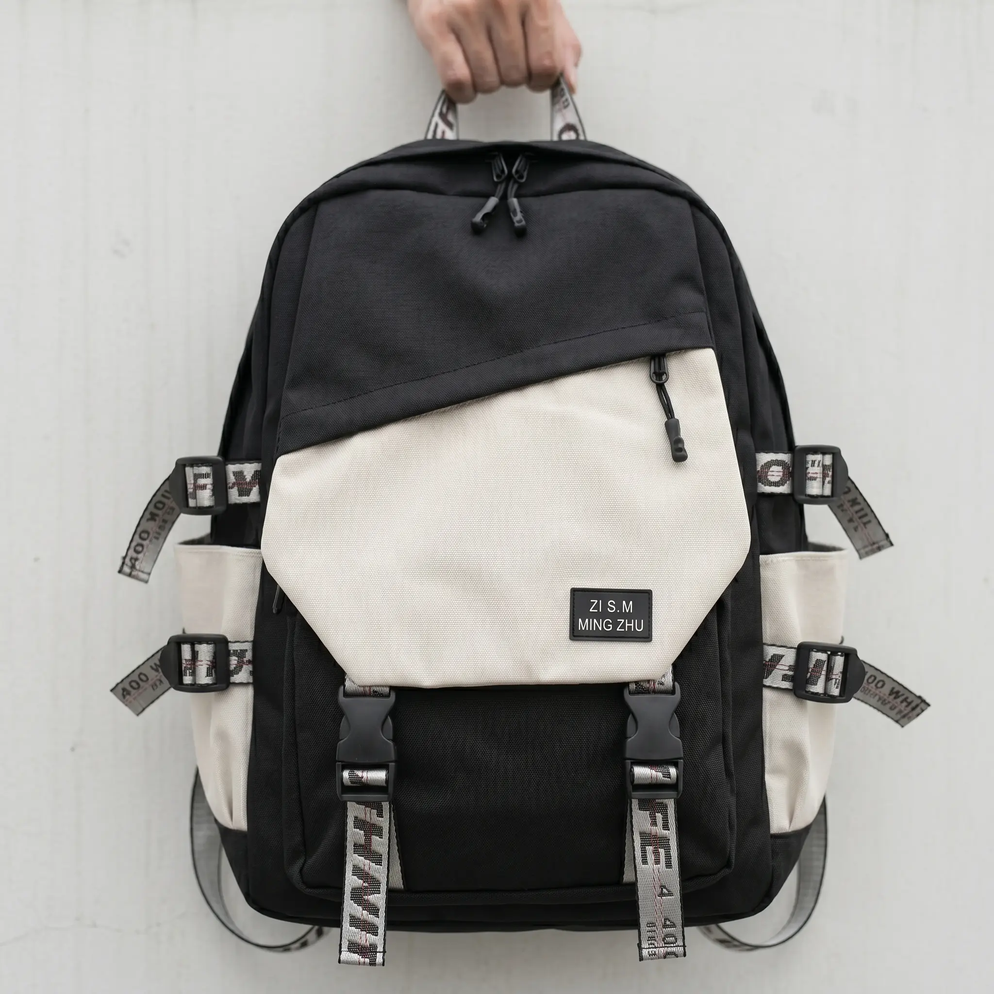 A person holding a durable black and beige contrast urban streetwear backpack by the top handle against a minimalist grey wall. The backpack features an asymmetric beige front zip pocket with a "ZI S.M MING ZHU" woven patch, side compression buckle straps with patterned industrial webbing, dual bottom clip straps, and cream side water bottle pockets.