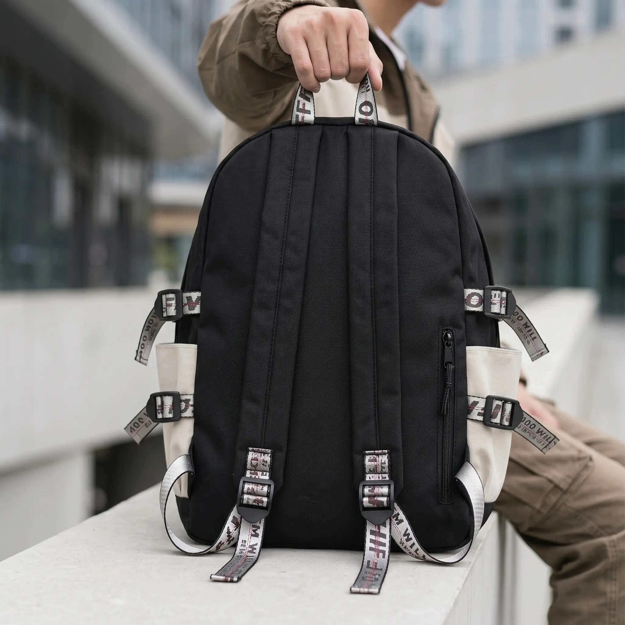 Rear view of a person holding the back of a black Tortokart urban utility backpack sitting outdoors. The image showcases the padded black shoulder straps with industrial webbed pull tabs, side compression buckled straps, cream side water bottle pockets, top carry handle, and a hidden vertical anti-theft zipper pocket on the lower right back panel.